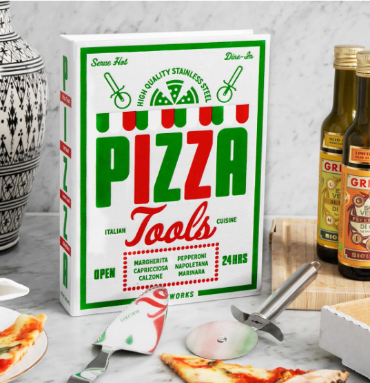 A boxed set of Pizza Tools kitchen utensils stands on a marble countertop with pizza slices, olive oil bottles, and cheese visible around it. The main product is the white box labeled 'PIZZA Tools' in large green and red lettering, with a pizza cutter and other utensils partly visible in front.