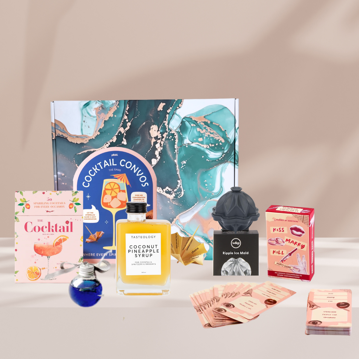 A curated gift set featuring a large marbled teal and gold cocktail-themed box, a bottle of coconut pineapple syrup, a pink cocktail book, a blue bottle, a black espresso machine, a pink lip gloss box, and several pink ticket-like accessories arranged on a light beige surface against a soft beige backdrop.