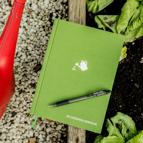 A green hardcover notebook titled 'MY GARDENING HANDBOOK' with a small white watering can icon on the cover, placed on a wooden garden stake with a black pen resting on it. The notebook is surrounded by gravel, soil, and green lettuce leaves, with a red watering can visible on the left.
